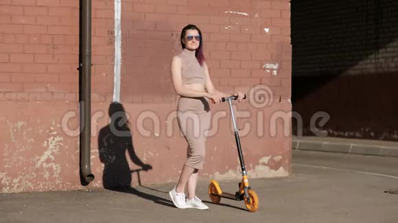 年轻漂亮的女人穿着裙子和太阳镜在砖墙的背景上骑着一辆踢踏板车兵马俑视频的预览图