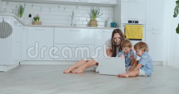 年轻漂亮的妈妈和孩子们坐在客厅的地板上看着笔记本电脑屏幕视频的预览图