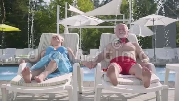 年长的积极夫妇躺在游泳池附近的日光浴床上手牵着手微笑充满爱意的家庭酒店休息视频的预览图