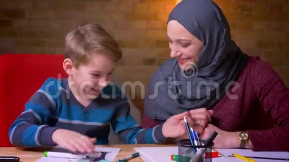快乐的穆斯林母亲戴着头巾帮助她的小儿子在舒适的家庭氛围中用标记画这幅画视频的预览图