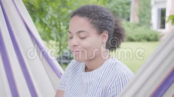 可爱的年轻微笑的非洲裔美国妇女坐在吊床上在花园里放松发短信的肖像视频的预览图