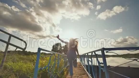 快乐的十七岁女孩穿着工作服举起双手在夕阳和湖面的背景下奔跑视频的预览图