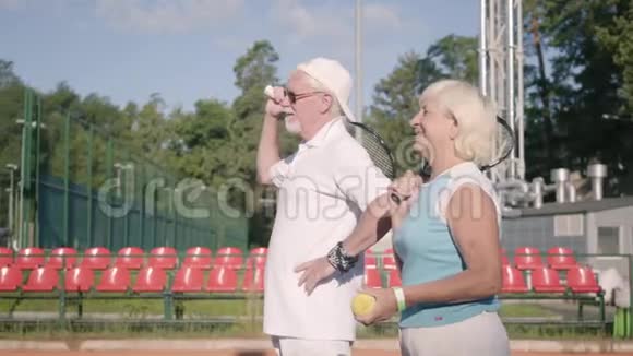 戴着太阳镜和网球拍的成熟夫妻站在阳光下的网球场上户外娱乐休闲视频的预览图