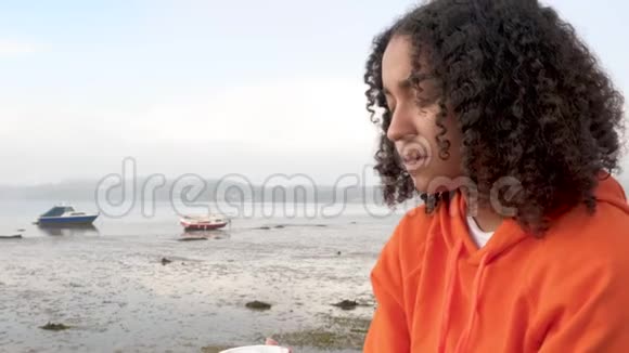 穿着橙色连帽衫的非裔美国少女少女在港口边悲伤或沮丧地喝着外卖咖啡视频的预览图