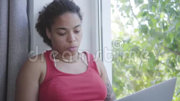 肖像可爱的年轻非洲裔美国妇女坐在窗台上在她的笔记本电脑上打字快乐女孩放松视频的预览图