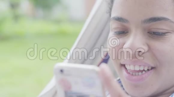 躺在吊床上在花园里放松用手机发短信的快乐非裔美国妇女的特写脸视频的预览图
