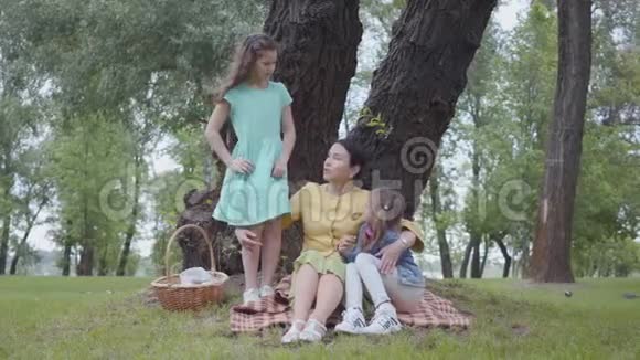 优雅的高级女人坐在公园树下的毯子上和两个可爱的孙女交谈一个女孩视频的预览图