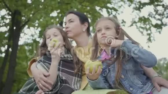成熟的女人和两个可爱的孙女坐在公园的树下吃苹果的女孩看着远处快乐视频的预览图