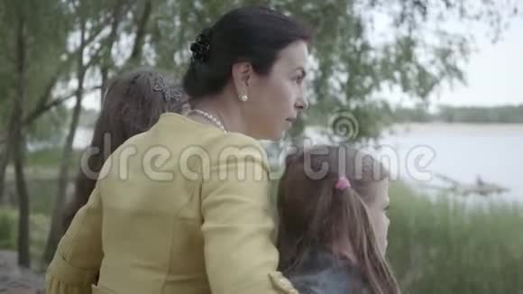 成熟的祖母拥抱她的两个孙女靠近美丽的河流快乐的家庭花时间在户外概念视频的预览图