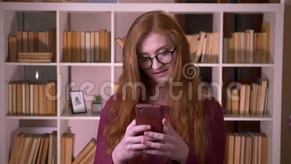 在大学里戴着眼镜的年轻红发美女戴着眼镜在手机上浏览的特写肖像视频的预览图