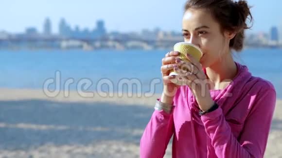 穿着粉红色运动服的美丽年轻运动女子在城市沙滩上用纸杯喝咖啡的画像视频的预览图