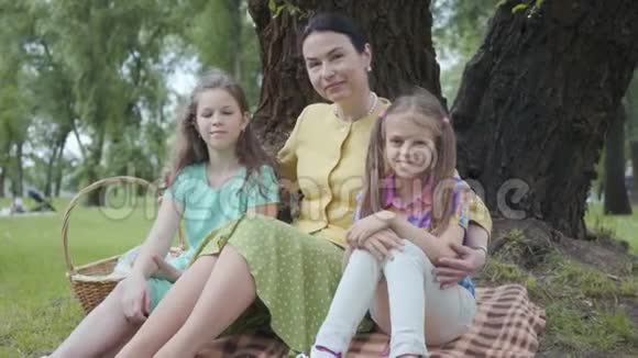 成熟的女人坐在公园树下的草地上和两个可爱的孙女一起看着镜头微笑着视频的预览图