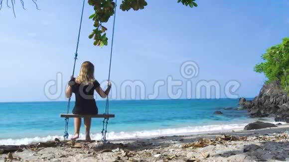 穿着黑色裙子的年轻女子在海滩上摇摆着蓝色的海洋景观快乐的女人在夏日海滩上荡秋千视频的预览图