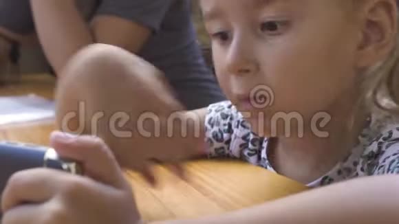 肖像可爱的小女孩坐在桌子旁看手机用智能手机闭脸搞笑女孩看视频的预览图