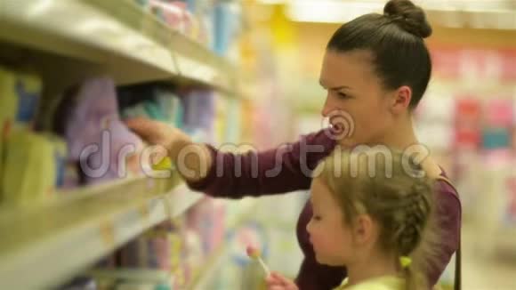 可爱的女孩和棒棒糖坐在超市手推车母亲在后台选择湿巾视频的预览图