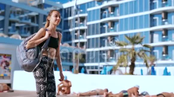 美丽苗条的年轻女子穿着运动服带着小背包在海岸上与大酒店对峙快乐的女人视频的预览图