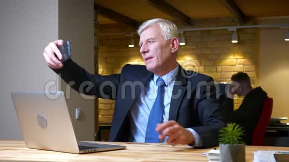 一位老白种人商人坐在笔记本电脑前的手机上自拍的特写镜头视频的预览图