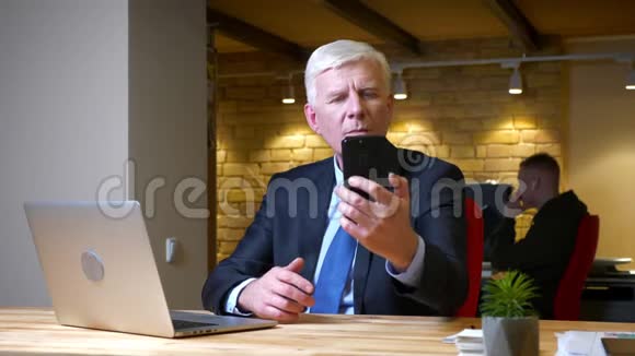 一位老白种人商人坐在笔记本电脑前的手机上自拍的特写镜头视频的预览图