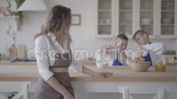 穿着围裙的漂亮女人站在前台看着她的两个儿子在厨房里快乐地做面团视频的预览图