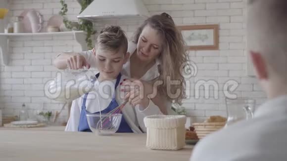 漂亮的女人和她十几岁的儿子在厨房做饭那个男孩帮妈妈做食物妈妈在碗里倒牛奶视频的预览图