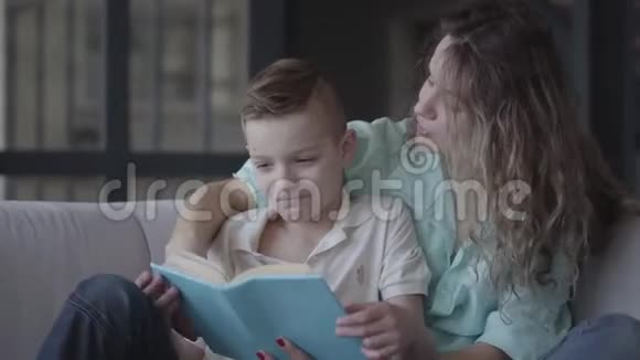 一位美丽的年轻母亲给坐在沙发上的漂亮儿子读一本书的肖像兄弟情谊家庭视频的预览图