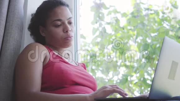 肖像可爱的年轻非洲裔美国妇女坐在窗台上在她的笔记本电脑上打字快乐的女孩在家放松视频的预览图