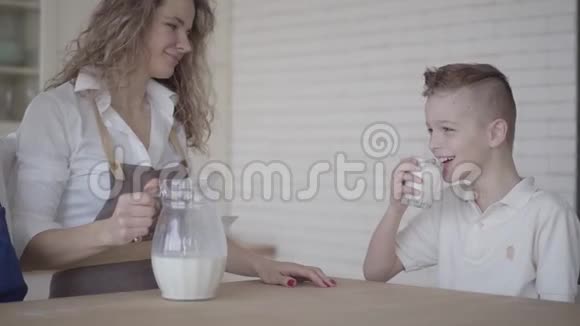 一个男孩在厨房里坐在他母亲和哥哥旁边的桌子旁喝牛奶兄弟情谊家庭视频的预览图