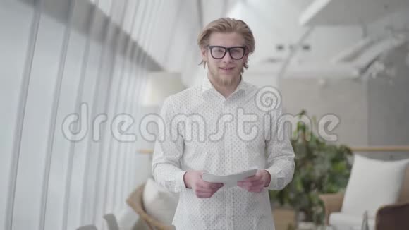 一位戴眼镜的成功金发男子站在一间光线舒适的办公室里拿着纸看着镜头视频的预览图