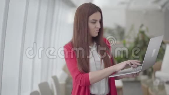 穿红色西装的年轻女子在现代光办公室的小工具上打字女孩合上上网本微笑着办公室视频的预览图