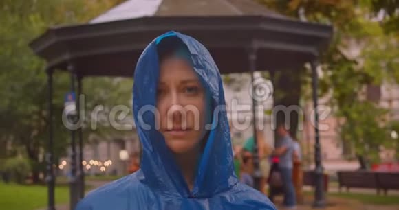穿着蓝色雨衣的白种人年轻女性在雨中站在绿色公园里看镜头的特写镜头视频的预览图