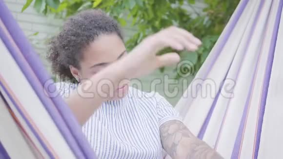 年轻微笑的非洲裔美国妇女坐在吊床上在花园里放松并发短信视频的预览图