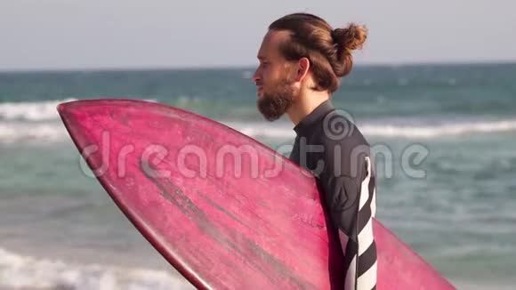 一位留着胡子的年轻时尚男士正拿着冲浪板站在海滩上视频的预览图