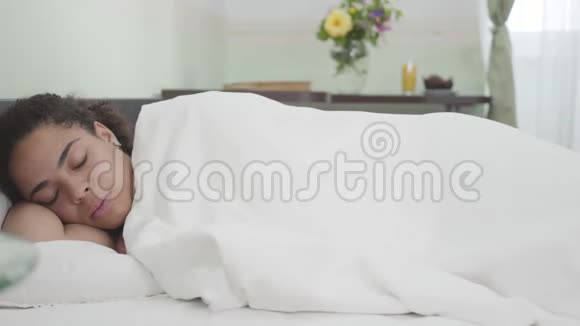 漂亮的非洲裔美国妇女睡在白色毯子下在灯光室的背景手机躺着视频的预览图