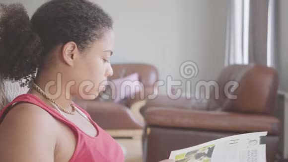 漂亮的非裔美国女人坐在扶手椅上咀嚼苹果看杂志特写快乐女孩放松视频的预览图