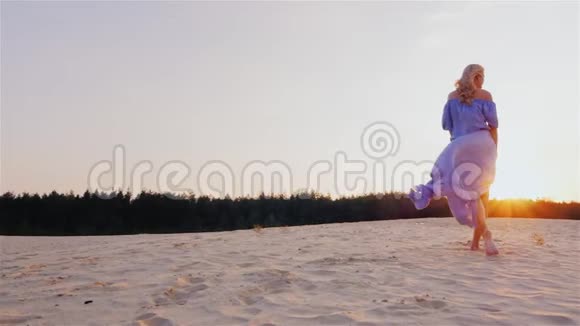 夕阳西下一个穿着浅蓝色连衣裙的苗条女人站在海滩上风吹着她的裙子视频的预览图