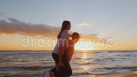 一对相爱的年轻夫妇在日落时在海滩上玩得很开心一个女孩坐在沙滩上奔跑的男人的肩膀上视频的预览图