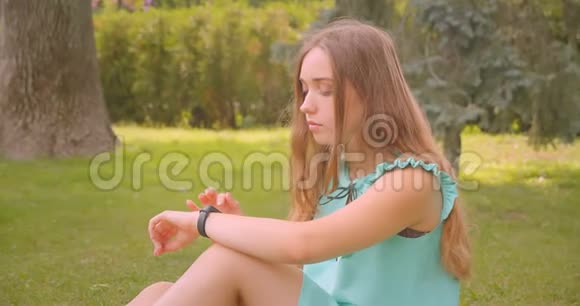 年轻可爱长毛美丽的女性坐在草地上用智能手表在公园户外休息视频的预览图