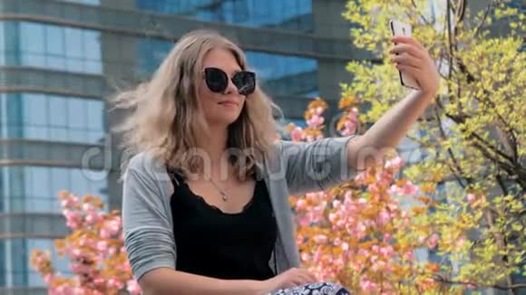 年轻漂亮的欧洲女孩戴着太阳镜坐在街上用智能手机自拍视频的预览图