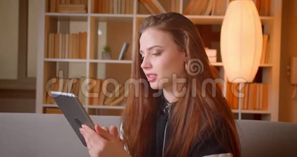 年轻的少女坐在书架上的沙发上坐在平板电脑里在家里用背景手表拍摄的画像视频的预览图