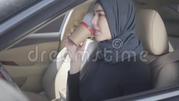 独立微笑的年轻穆斯林妇女坐在车里喝咖啡戴着电话交谈的肖像视频的预览图