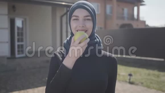 独立的年轻穆斯林妇女穿着传统头巾咬着多汁的绿苹果的肖像视频的预览图