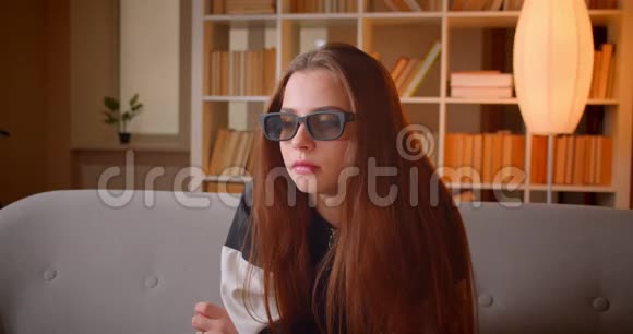 年轻少女戴着3D眼镜的肖像坐在沙发上看电视坐在书架背景上看电影视频的预览图