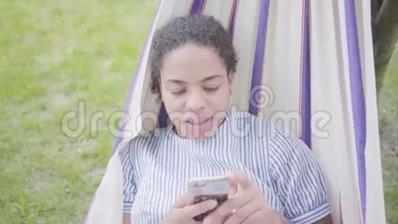 躺在吊床上在花园里放松在手机上发短信的美国年轻非洲裔妇女的肖像视频的预览图