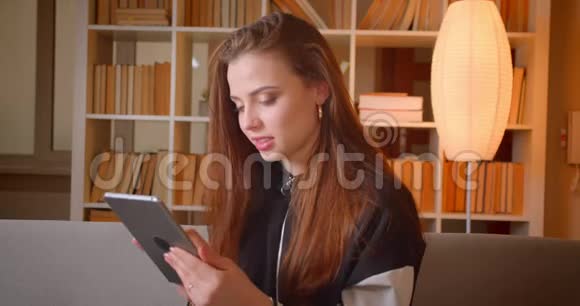 年轻的少女在书架上坐在沙发上用平板电脑工作的肖像背景手表在家里变成相机视频的预览图