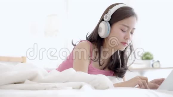 美丽的年轻亚洲女人躺在卧室里用手提电脑放松听音乐视频的预览图