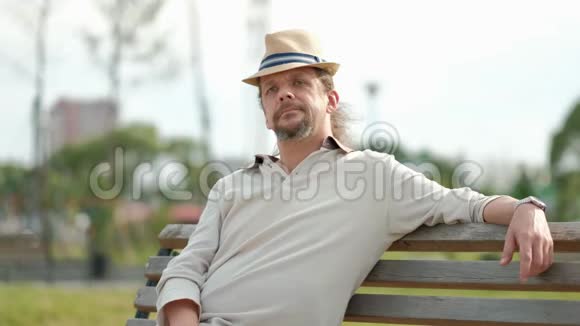 放松迷人的中年男子长发戴着帽子坐在公园的长凳上休息夏季傍晚炎热视频的预览图