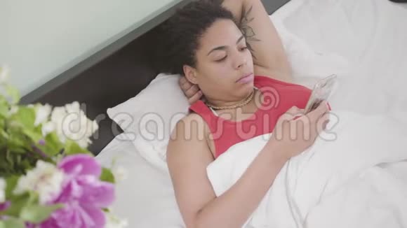 年轻的非裔美国女人刚醒来她躺在床上用手机发短信快乐的女孩在家放松日视频的预览图