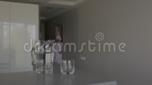 一个穿着白色长袍头上有一条毛巾的女人把水壶里的水倒进玻璃杯里喝视频的预览图
