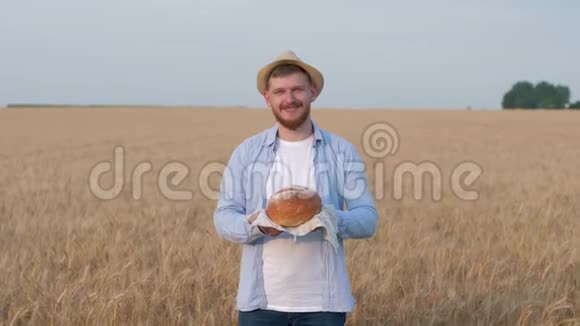 面包师肖像戴草帽的年轻人给你新鲜出炉的面包在麦田里对着镜头微笑视频的预览图