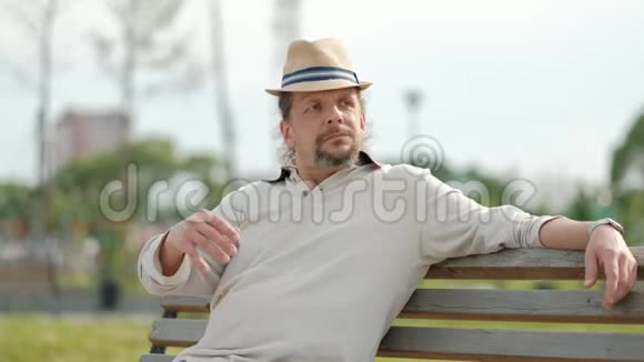 放松迷人的中年男子长发戴着帽子坐在公园的长凳上休息夏季傍晚炎热视频的预览图